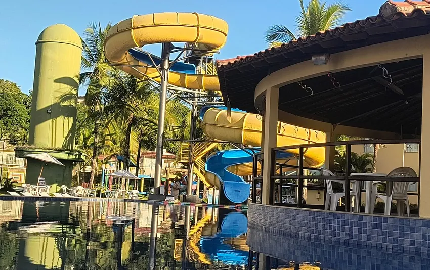Foto da área de lazer de um resort mostrando uma piscina, um grande toboágua caracol amarelo e azul, coqueiros e um quiosque