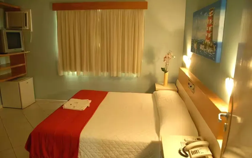 Foto de um quarto com cama de casal, janela ao fundo e frigobar no canto